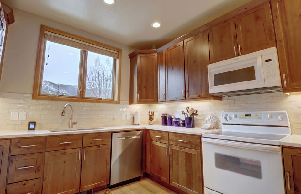 A modern kitchen with wood cabinets, white appliances, and a window above the sink. The countertop hosts a knife set and a blue utensil holder next to the stove.