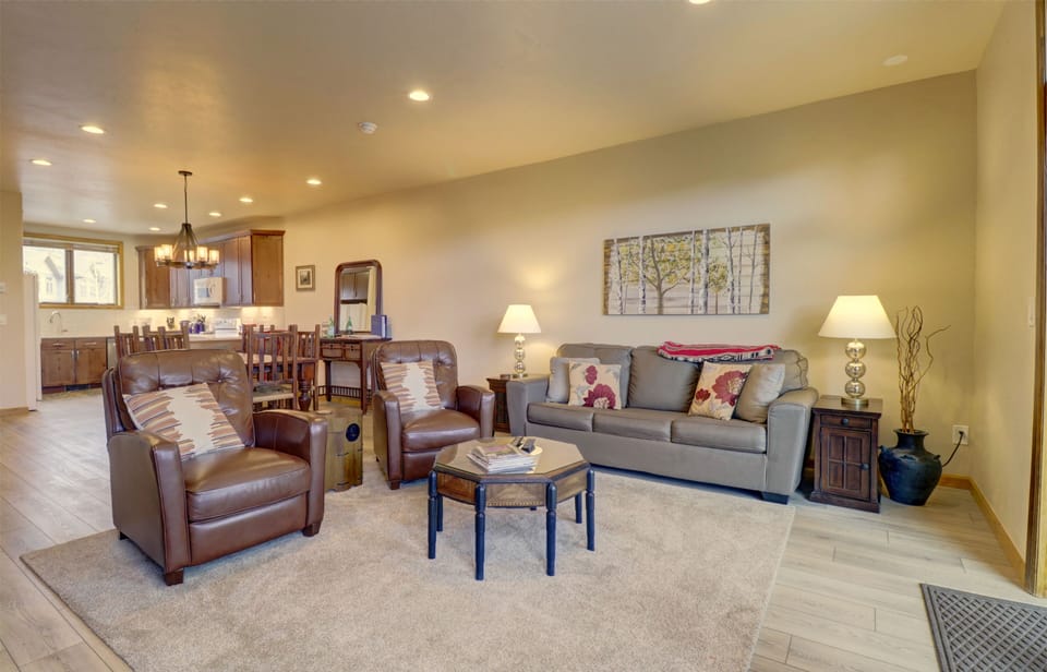 A cozy living room with a sofa, two leather armchairs, and a coffee table. The room features warm lighting, wooden flooring, a dining area in the background, and a wall painting above the sofa.