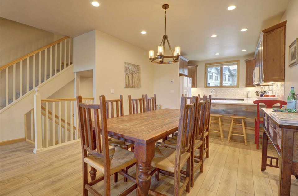 A rustic dining room with a wooden table and eight chairs. The room features a staircase, chandelier, and a kitchen area with wooden cabinets and modern appliances. Natural light streams through windows.