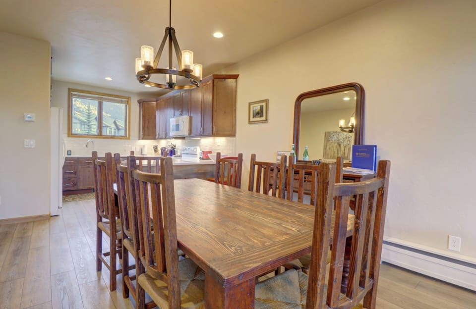 A dining area with a wooden table and eight chairs, a chandelier above, and a kitchen with wooden cabinets in the background. A large mirror and a few framed pictures are on the walls.