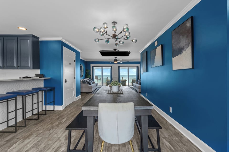 Gather around the dining table beneath a bold chandelier, where ocean views, rich blue walls, and modern design create a coastal space that’s equal parts stylish and welcoming