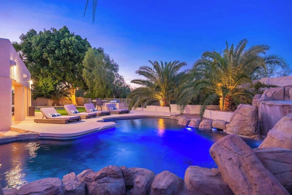 Luxurious resort-style pool features a dramatic waterfall flowing over natural rock formations. Palm trees frame the illuminated waters at twilight, while poolside loungers await under desert skies.