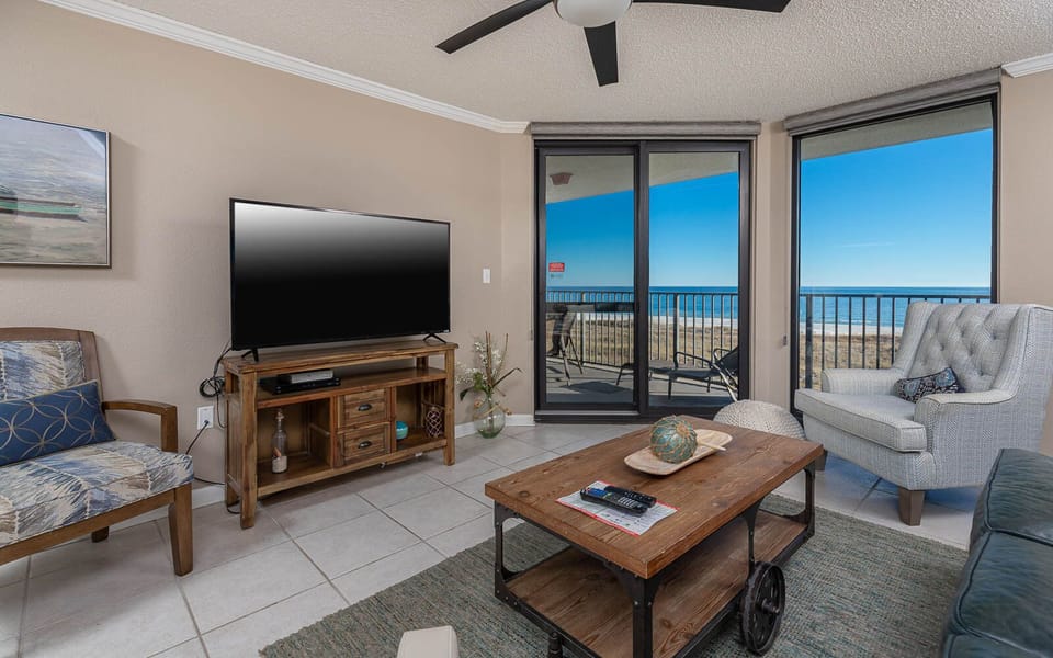 Living area features a cozy sofa, accent chairs, and a large TV, with sliding doors revealing a beautiful beachfront view