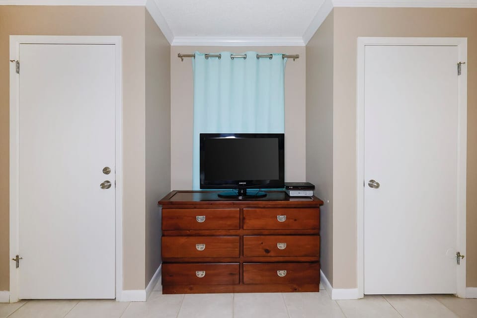 Wooden dresser with six drawers and a TV on top, flanked by two doors and accented by a light blue curtain in the guest room