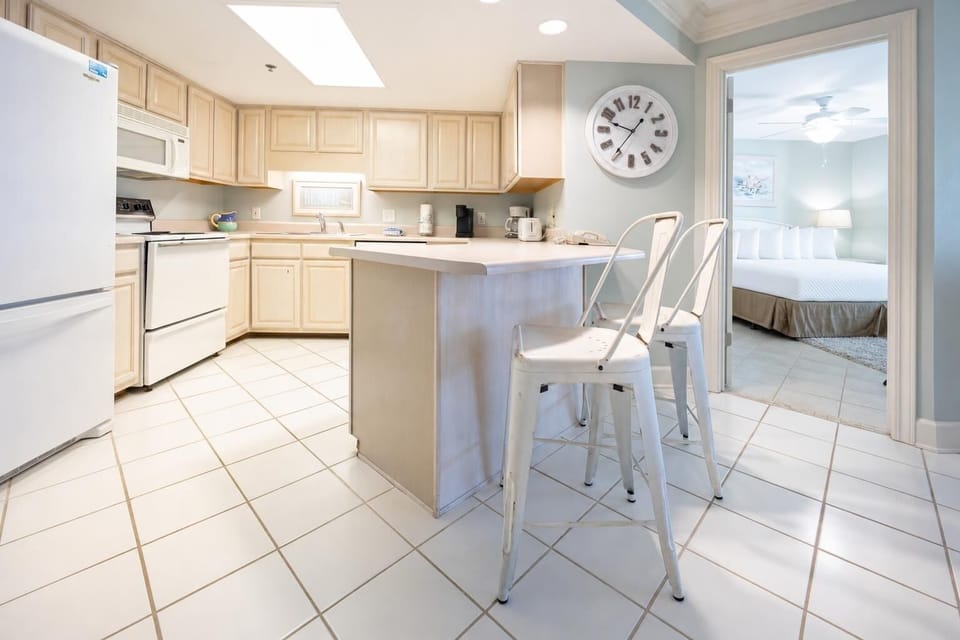 Bright kitchen area features light cabinetry, a breakfast bar with seating, and a view into a cozy master suite beyond