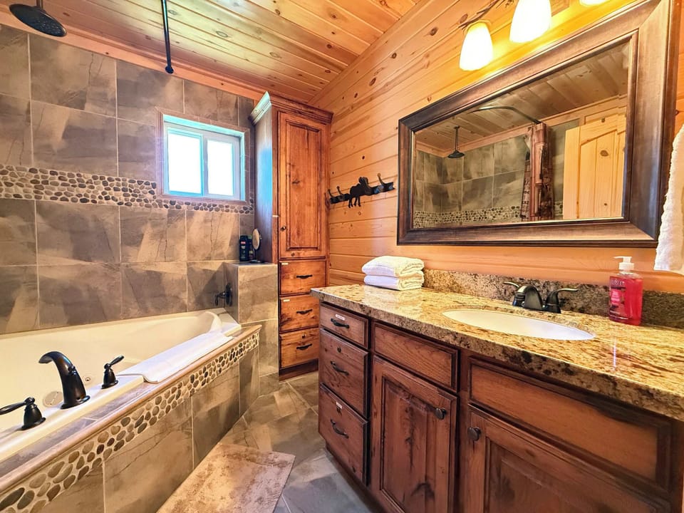 The luxury en-suite master bathroom boasts a large custom wooden vanity with a premium granite countertop, bright lighting, and a deep soaking jetted tub.