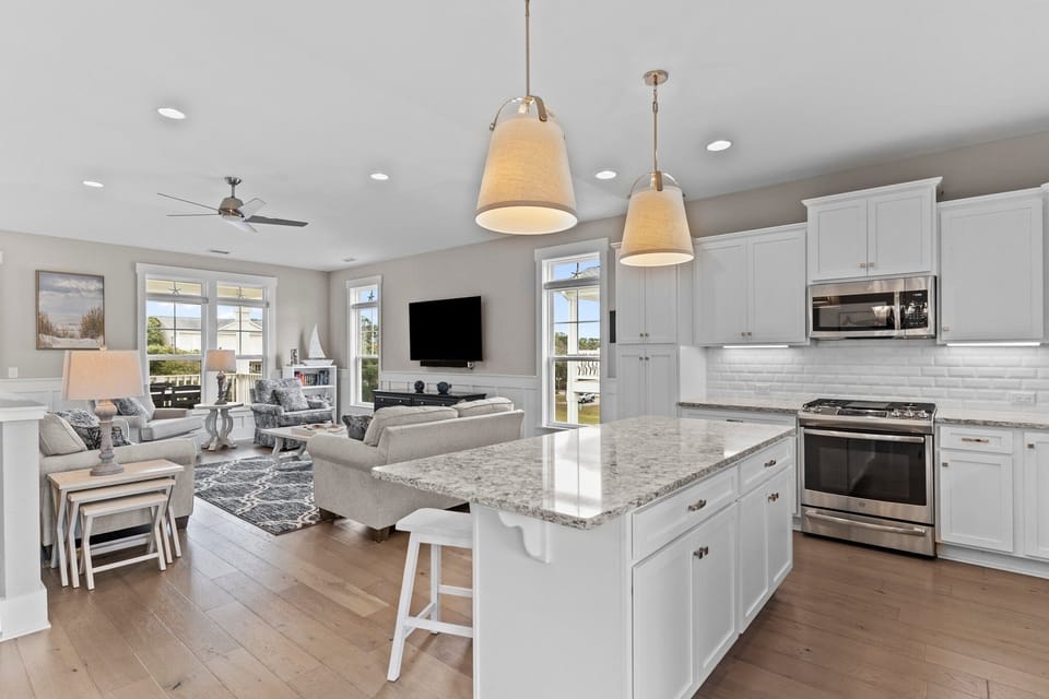 Bright coastal kitchen with white cabinetry, granite countertops, subway tile backsplash, stainless appliances, large island with seating, pendant lighting, and window over the sink.