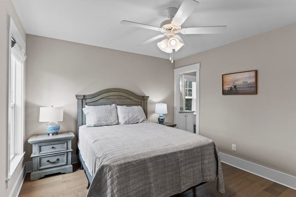 Relaxing coastal bedroom with queen bed, nightstands with lamps, ceiling fan, hardwood floors, and private bathroom access. Bright, comfortable space perfect for unwinding after an Oak Island beach day.