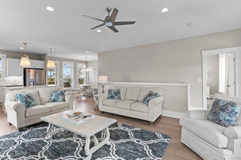 Light-filled living room with plush sofa, patterned armchairs, coastal décor, coffee table, area rug, ceiling fan, and large windows with door opening to the covered balcony.