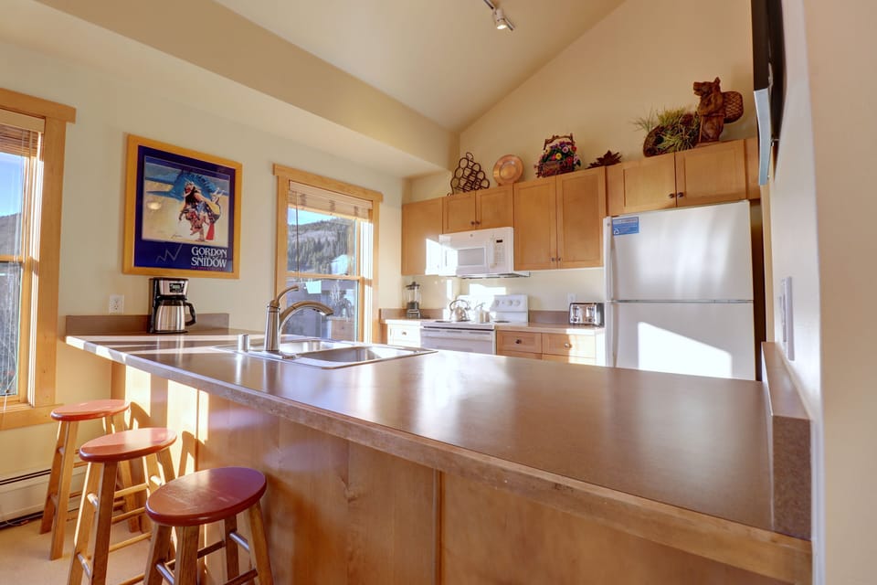 A kitchen with light wood cabinets, a white refrigerator, stove, and microwave, a double sink, and a breakfast bar with three wooden stools. A window and wall art are visible.
