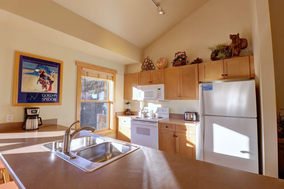 A kitchen with wooden cabinets, a white microwave, stove, and refrigerator. The counter has a sink and a coffee maker. A window and framed poster are on the wall. Various decorations are above the cabinets.