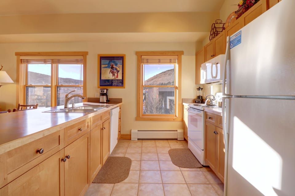 A bright kitchen with light wooden cabinets, white appliances including a fridge and a microwave, a central island with a sink, and two windows revealing a mountainous landscape.