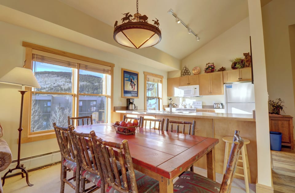 A well-lit kitchen and dining area with a rustic wooden dining table, chairs, a round chandelier, and large windows. The kitchen has wooden cabinets, white appliances, and various decorations.