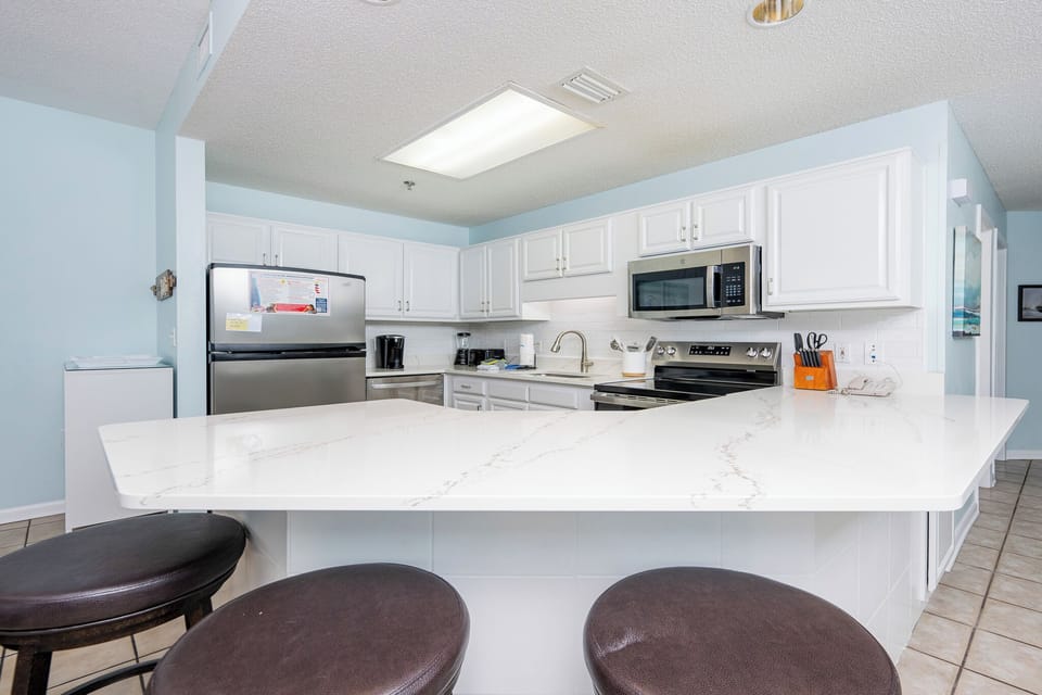 The kitchen features white cabinetry, coffee station, a spacious breakfast bar with seating, and stainless appliances