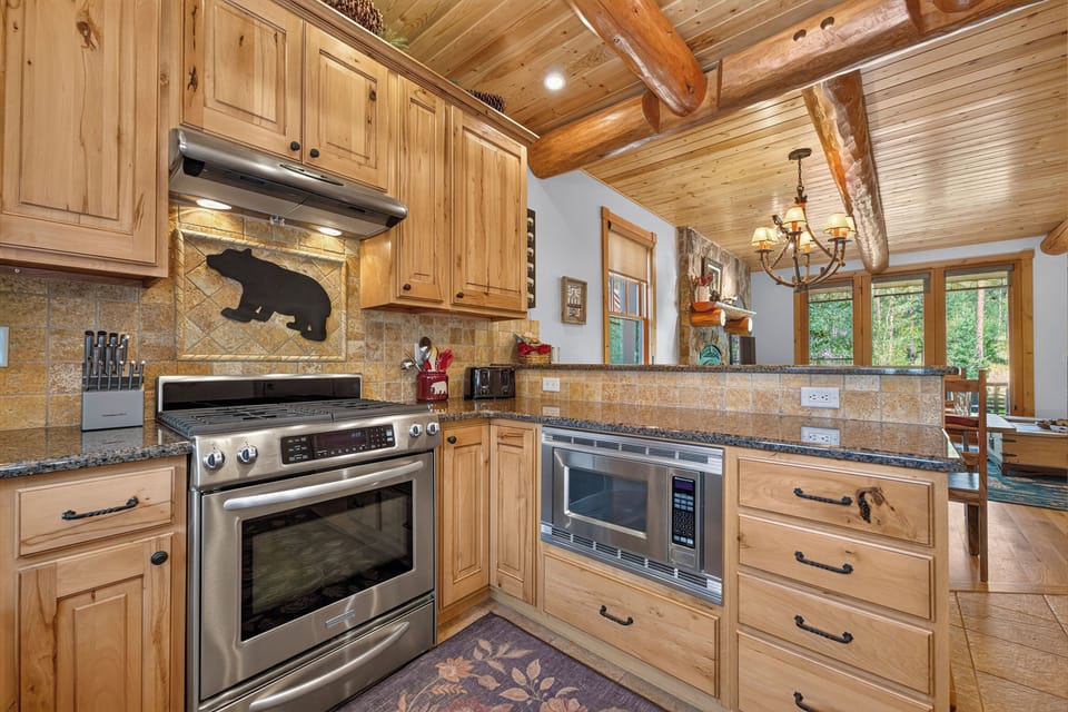 Rustic mountain kitchen featuring warm wood cabinets, granite counters, and stainless-steel appliances. Charming bear silhouette and exposed log beams create authentic cabin character.
