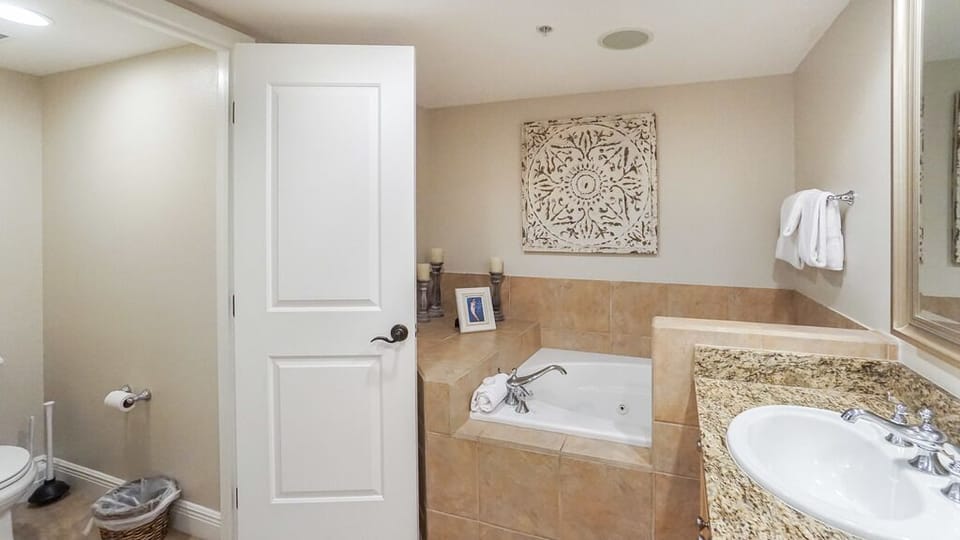 Step into your private spa-like bathroom featuring a deep soaking tub framed in warm travertine tiles. Fresh towels await your arrival, and the elegant wall art adds a touch of coastal charm.