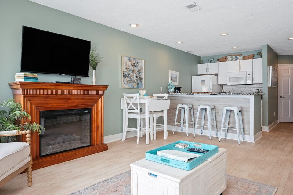 Modern open-concept living space featuring a warm wood fireplace, soft sage walls, and a bright kitchen breakfast bar. 🏡🌊