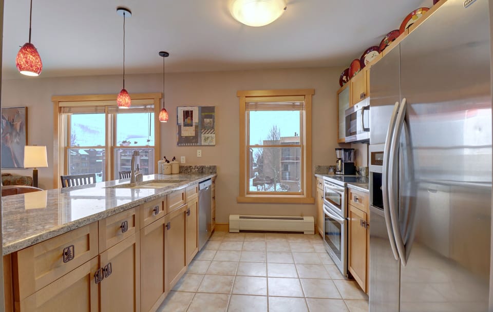 Modern kitchen with stainless steel appliances, granite countertops, wooden cabinets, and tiled floors. Red pendant lights hang above the island. Large windows offer natural light and a view outside.