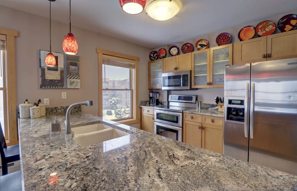 A modern kitchen features a stainless steel fridge, oven, and microwave, wooden cabinets, and a granite countertop island with a sink. Decorative plates are displayed atop the cabinets.