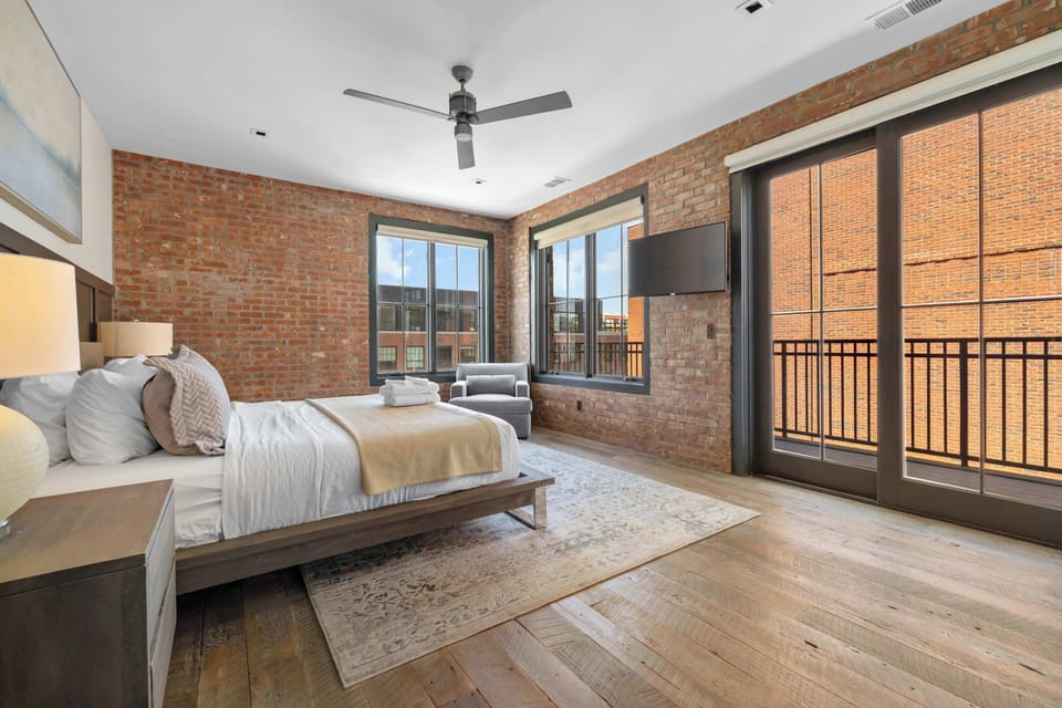 Bright bedroom angle showcasing large windows, hardwood floors, and a modern ceiling fan.