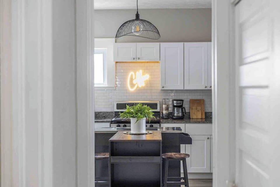 Charming kitchen with a central island featuring rustic stools and a leafy centerpiece. Behind, a neon “Café” sign glows above a sleek stove, flanked by white cabinets and a cozy coffee station. ️☕