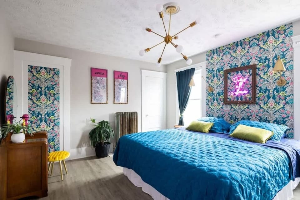 Vibrant bedroom featuring a queen-sized bed dressed in bright blue linens and green accent pillows against a floral wallpapered accent wall. Gold fixtures and natural light create a stylish, cheerful retreat. ️-2nd Floor