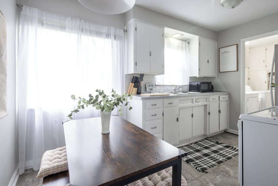 - Bright white kitchen with dining table and cushioned seating   - Modern Airbnb stay with natural light and homey details   - Functional cooking space perfect for family meals 