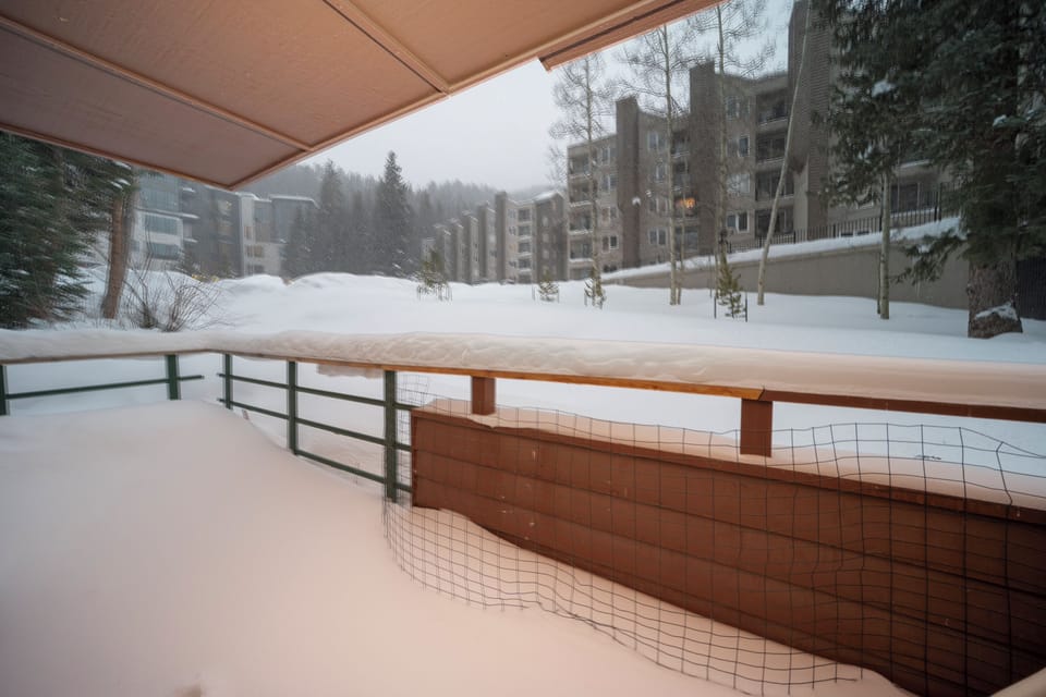 Snow-covered terrace overlooking a peaceful winter resort setting with mountain buildings and pine trees.