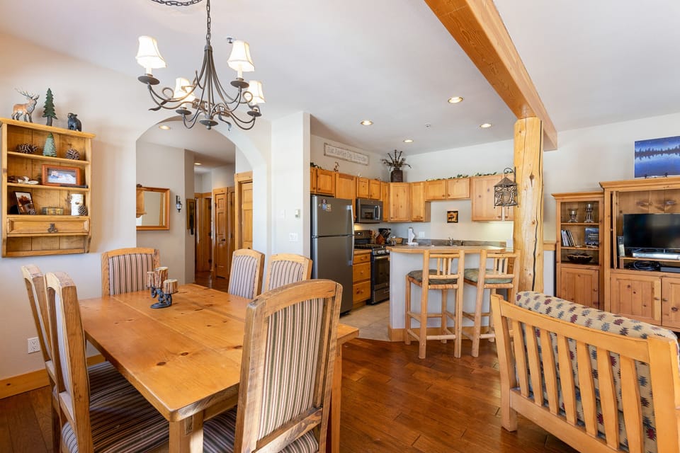 A cozy, rustic kitchen and dining area with wooden furniture, a chandelier, and visible decor items. The kitchen features an island counter with bar stools, and the dining table has six chairs.