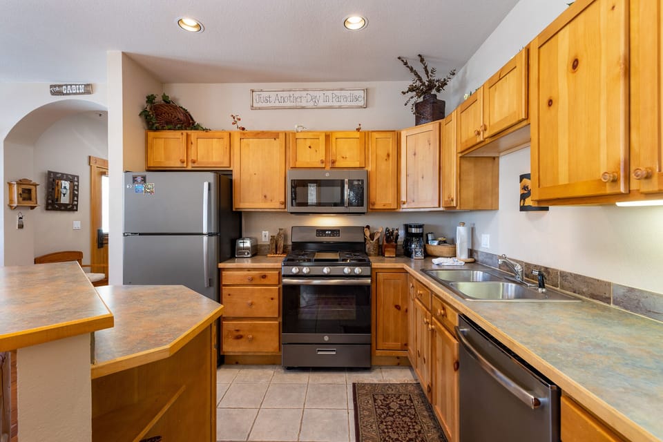A kitchen with wooden cabinets, stainless steel appliances, and a sign above the stove reading "Just Another Day in Paradise." The countertop is L-shaped, and there is a double sink on the right.