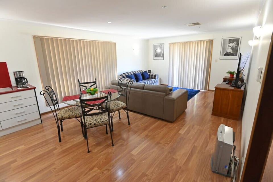 This bright open-plan space features a cozy living area with a grey sectional sofa and a checkered armchair, accented by a plush blue rug. A round glass dining table with four cushioned chairs sits nearby, adjacent to the modern kitchen. Vertical blinds a
