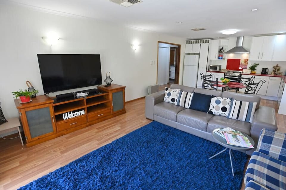 This open-plan living area features a cozy lounge with a large TV on a wooden entertainment unit, a modern grey sofa adorned with patterned cushions, and a vibrant blue rug. The space flows seamlessly into a fully-equipped kitchen with white cabinetry, a 