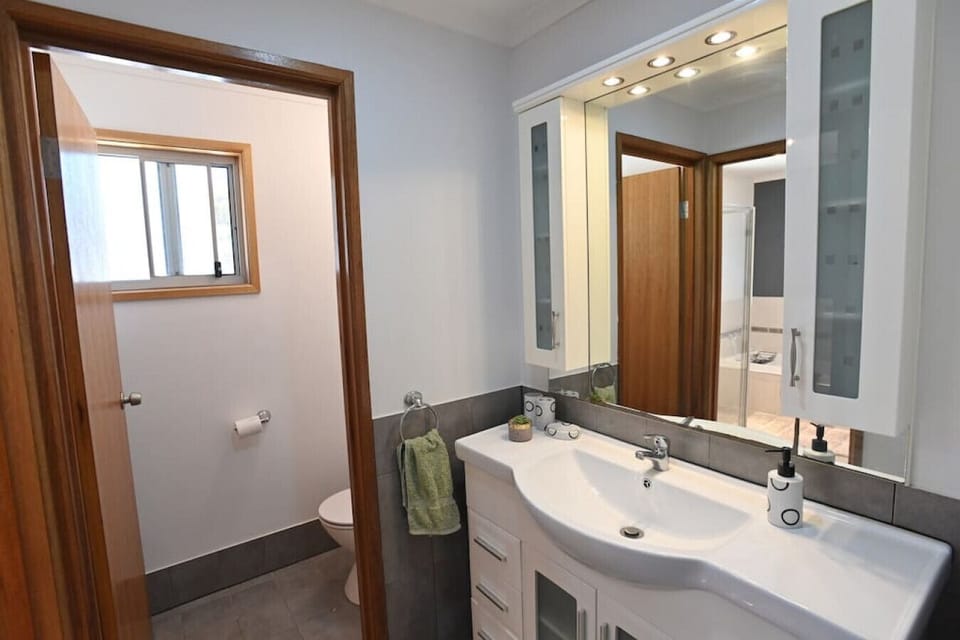 This modern bathroom features a sleek white vanity with ample drawer storage and a large mirror flanked by illuminated cabinets. A separate toilet area with a frosted glass window provides privacy, while the grey-tiled floor and neutral walls give the spa