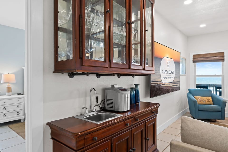 Your elegant wet bar with gleaming glassware awaits, perfect for crafting drinks while ocean breezes drift through nearby windows and soft natural light fills the space.