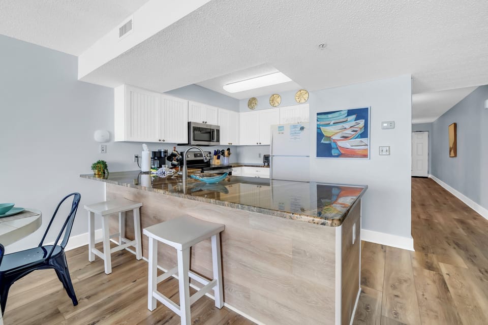 Gather around this bright kitchen island where granite countertops reflect coastal artwork, creating your perfect space for morning coffee and evening conversations.