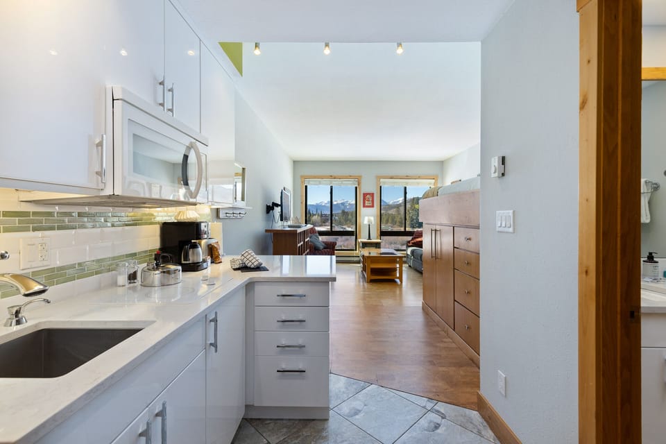 A modern, compact studio apartment with a white kitchen, light-colored walls, wooden furniture, and large windows offering a mountain view.