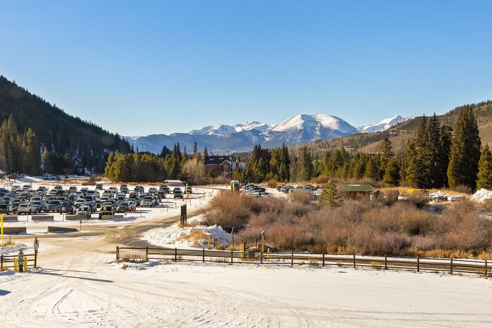 A large parking lot filled with cars sits near a snow-covered area and a mountain range under a clear blue sky. Trees and buildings are visible in the background.