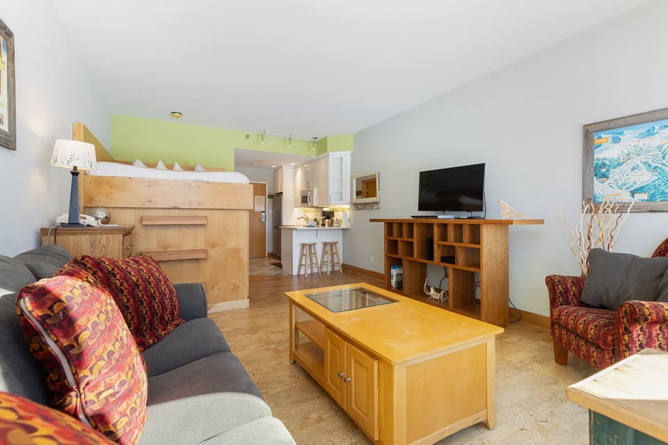 A living room with a couch, armchair, coffee table, wall-mounted TV, and lofted bed in the background. The room has wooden furniture, light-colored walls, and a glimpse of a kitchen area.