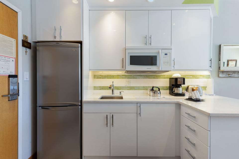 Modern kitchen area featuring white cabinets, a stainless steel refrigerator, a microwave, a coffee maker, and a small sink with a faucet. The countertop has a paper towel holder and a dish rack.