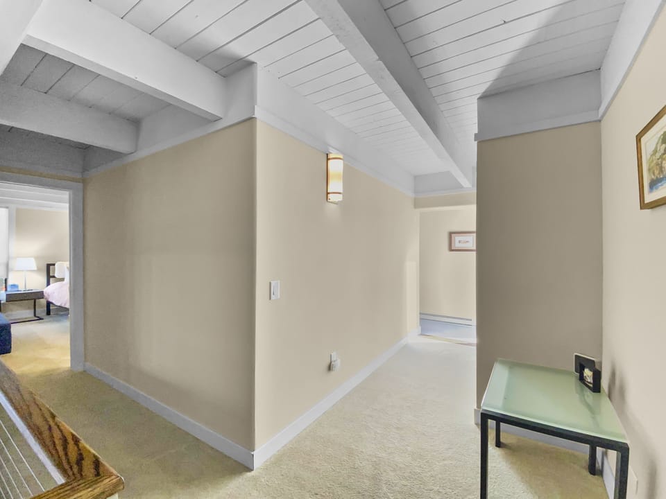 A hallway with beige walls and a white wooden ceiling, featuring a light fixture, a glass-topped table with a painting above it, and carpeted flooring, leading to a bedroom.