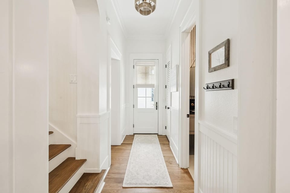 Narrow hallway with white beadboard walls, wooden stairs on the left, and a runner rug leading to a back door.