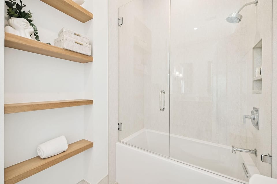 Bathroom featuring a white soaking tub with glass shower door and light wood floating shelves with decor and towel.