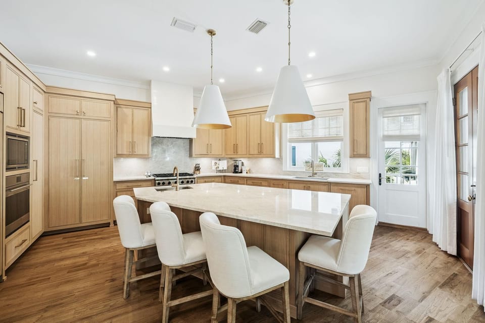 Light wood cabinetry surrounds the large marble island with white cushioned stools and pendant lighting above.