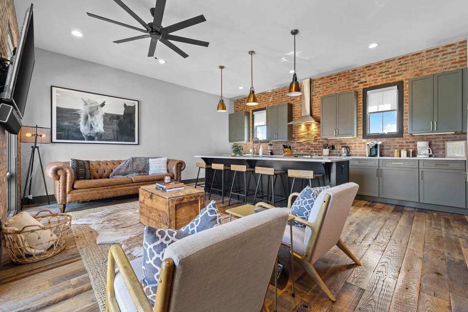 Bright and stylish living area featuring a cozy industrial design with exposed brick walls, large windows with plenty of natural light, comfortable seating, and a Smart TV
