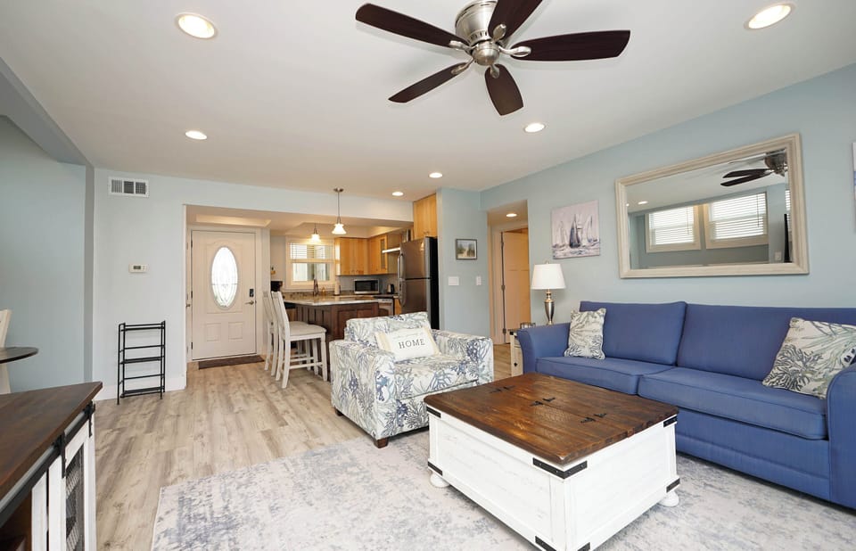 Relax in your bright, coastal-inspired living room with plush seating, ceiling fan, and open layout flowing to the kitchen.