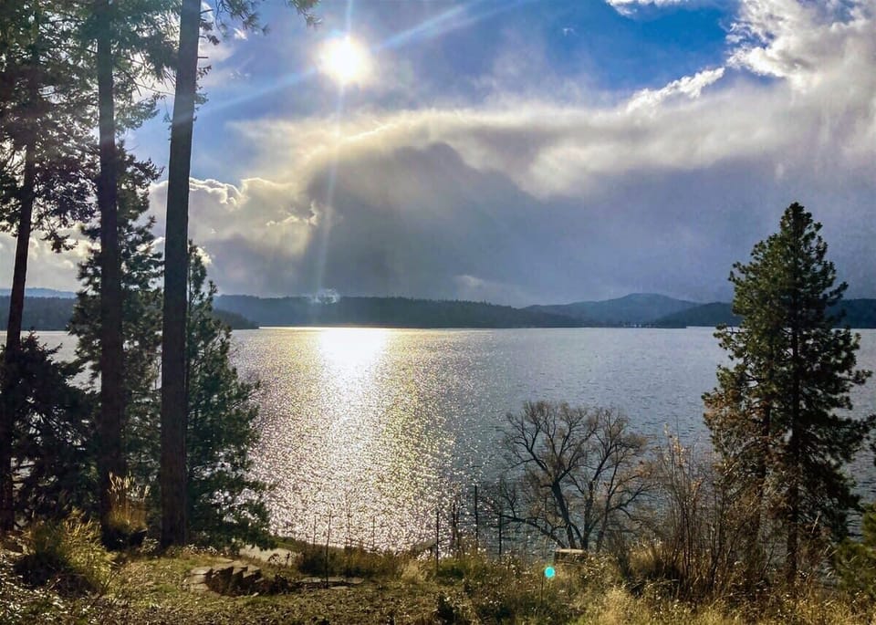 Pristine lake view framed by towering trees, with dramatic clouds casting golden light across the water and rolling hills beyond.