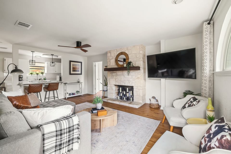 A large TV is mounted to the side of the fireplace, blending functionality with style. Two white, mid-century modern armchairs sit in front of a large, arched window. 