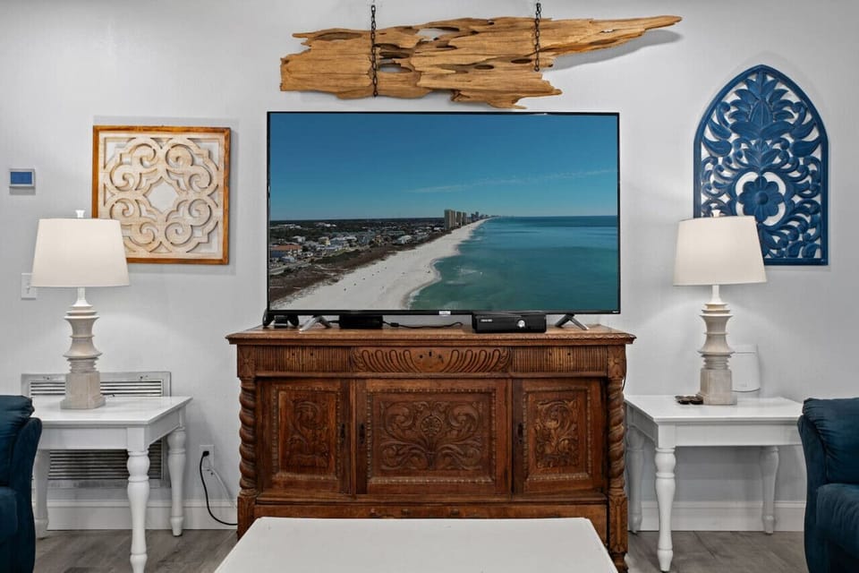 A large smart TV on a wooden brown dresser with intricate designs, surrounded by beautiful wall decor, and flanked by two smaller dressers with sleek white modern lamps on each.