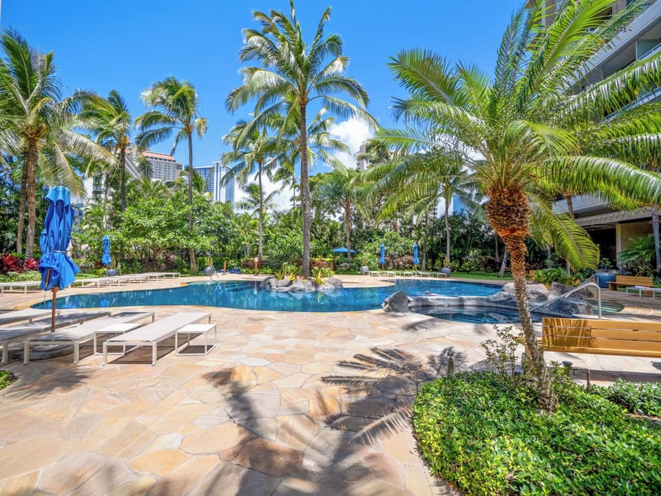 Pool and Hot Tub - A wider view shows the shared free-form pool, stone deck, lounge chairs, umbrellas, and surrounding palms, highlighting the landscaped recreation area available to guests of The Watermark Waikiki on extended stays.