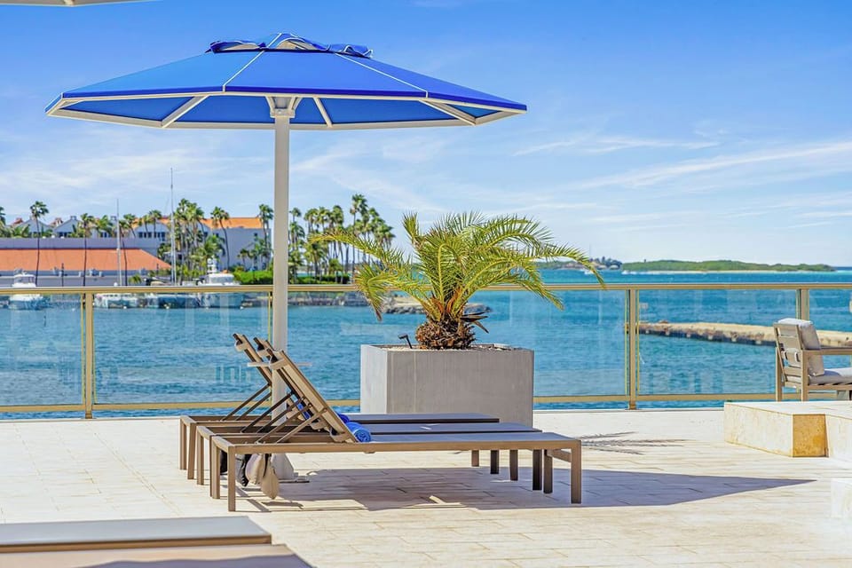Modern apartment complex featuring a striking glass-walled pool in an inner courtyard. The pool area is surrounded by a well-maintained landscaped hedge, vibrant blue umbrellas, and loungers—ideal for relaxation under sunny skies.